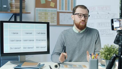 Online tutor recording video lesson using smartphone camera in classroom. Young man speaking explaining English grammar and gesturing.