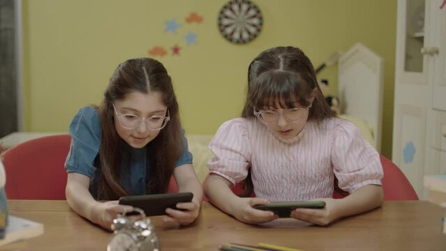 Two little girl friends playing online games with smartphones at home daycare. Children internet addiction. Portrait of girls playing games with smartphones.