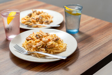 Two plates on the edge of a wooden table. Pasta with mussels and shrimp, water with lemon.