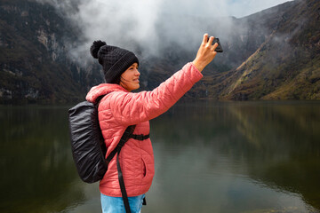 Chica sonriente con una chaqueta rosa parada junto al lago en las montañas y tomando selfies - obrazy, fototapety, plakaty