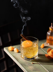 A glass of whiskey with ice garnished with tangerines and smoking cinnamon stick on wooden table.Close up of drink.
