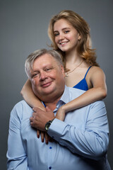 Dad and teenage daughter hug and smile. A girl in a blue dress, a man in a blue shirt. Love and tenderness. Grey background. Vertical.