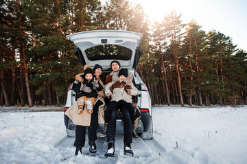 Happy young family having fun together. Parents sit with children in the trunk of car in winter forest. Family on winter walk. - obrazy, fototapety, plakaty