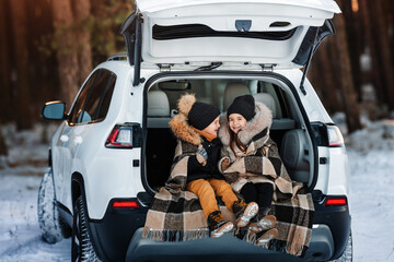 Two happy little children are having fun on a winter walk. Children sit in trunk of family car. Children warm themselves wrapped in warm blanket.