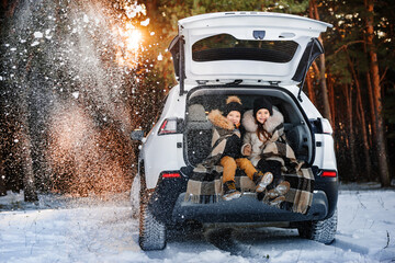 Two happy children on walk in winter forest. Children sit in trunk of family car. Children warm...