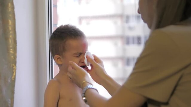 Crying baby. A caring mother applying antiseptic cream to a scratch and a bump on her 2-year old son's forehead and nose. A little boy was running on a playground on a hot summer day and fell.