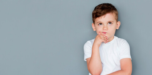 Emotional thinking little boy in casual outfit on gray background