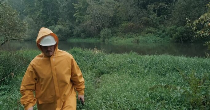 Construction engineer in protective helmet and yellow raincoat walks to river through wet, tall grass, man has phone in his hand. Concept of building bridges, cleaning banks and riverbeds of pollution