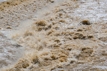 Rapids on the river after heavy rain in rainy season. Muddy water moving with great swiftness passing over a weir, falling down, splashing and forming bubbles.