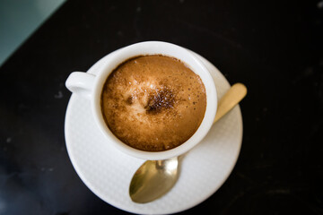 Porcelain white coffee cup with cappuccino, mochaccino on a saucer with a teaspoon on a black marble table to view. Hot tasty drink for a breakfast, branch, lunch. Aromatic beverage on a kitchen.