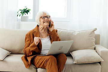 a sad, angry elderly woman in a brown suit is sitting with a laptop on her lap on a cozy sofa working from home and talking on the phone, looking serious and holding her hand in her fist