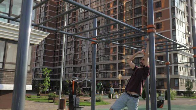 A bald with beard thin skinny guy is engaged on the crossbar on the street. A man training on a sports ground in a beautiful city park in casual clothes. Street workout, sport lifestyle.