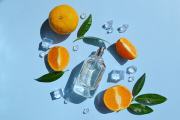 Glass bottle of perfume with oranges, green leaves, ice cubes, water drops on the light blue background with day sunlight. Smell of summer. Fresh orange cooling perfumery.  Direct sun rays. 