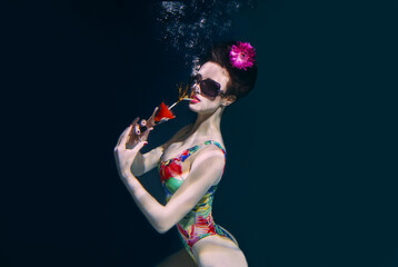 beautiful woman in a colourful stylish swimsuit and sunglasses and pink gerbera in the head with a cocktail in her hand underwater in the swimming pool
