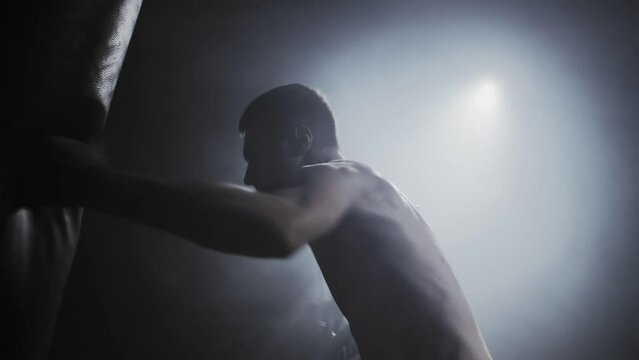 Portrait of a caucasian male boxer beating a punching bag in dark gym. Silhouette of a fighter training, light source in the background. Concept of motivation, willpower and fighting spirit