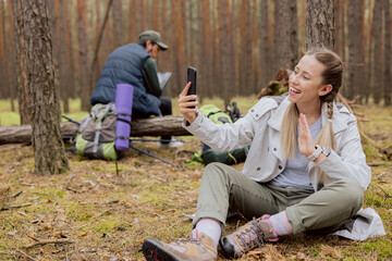 Couples in the woods smiling woman in the foreground with blonde hair tied in braids sits on the ground in the forest and talks on the phone video conversation, resting while trekking, camping, hiking
