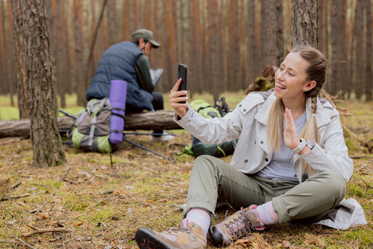 Couples in the woods smiling woman in the foreground with blonde hair tied in braids sits on the ground in the forest and talks on the phone video conversation, resting while trekking, camping, hiking