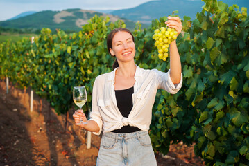Happy girl holding a bunch of grapes and a glass of white wine on the background of a vineyard