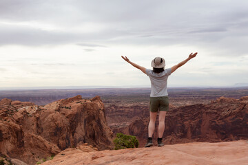 Adventurous Woman Hiking at a Desert Canyon with Red Rock Mountains. Cloudy Sky. Canyonlands National Park. Utah, United States. Adventure Travel