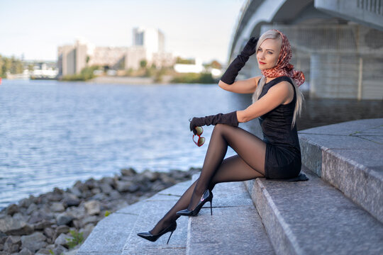 Beautiful blonde girl in black dress with perfect legs in pantyhose and shoes with high heels posing outdoor near the river bridge in the rays of sun. Street fashion photo.