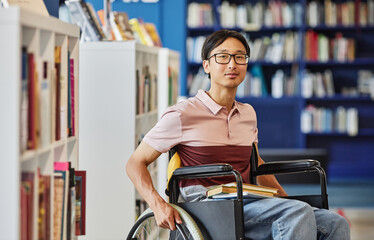 Vibrant portrait of Asian young man in wheelchair looking at camera in college library and smiling