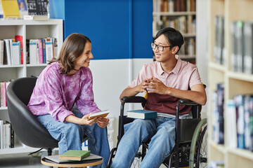 Portrait of smiling young man in wheelchair talking to friend while studying together in library