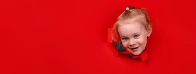 Portrait of young smiling girl looking through hole in the red paper wall, panoramic layout