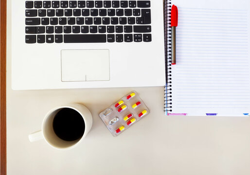 Simple clean image showing white laptop, pen, notebook, a cup with coffee and medicine on the table with space to write.