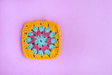 Closeup of multicolored bright crochet motif of square shape on light violet background.