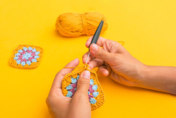 Closeup of woman's hands with a hook inserted in crochet element with the backgound of yellow table, yellow skein of yarn and finished granny square element - obrazy, fototapety, plakaty
