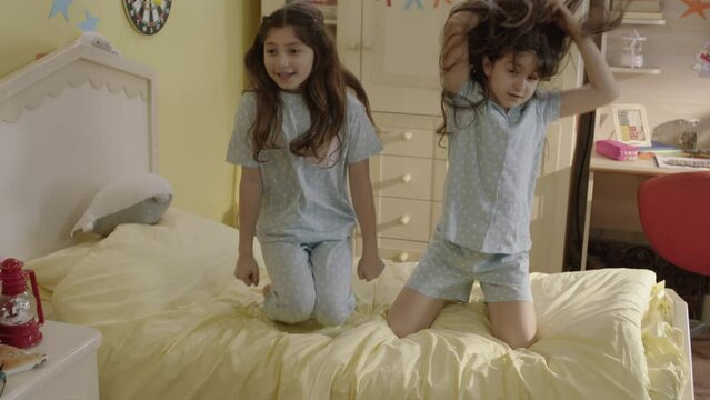 Two little girls in pajamas have fun jumping on the bed. Fun,cute happy child girls jumping on the bed,jumping, laughing,dancing to music.In the absence of their mother,the girl throws a pajama party.