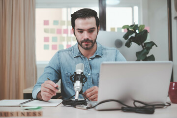 Young man host sitting at table, streaming audio podcast using microphone and laptop in studio, Social media, podcasting, blogging concept. Cheerful casual caucasian employee.