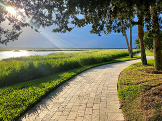 Brick path along a saltwater marsh glowing from sunbeams through tree branches.