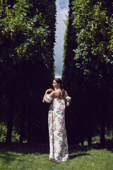 Outdoor portrait of a beautiful luxury brunette woman in a dress with flowers stands in a park with trimmed trees.