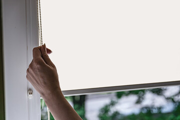 Closeup of woman hand opens roll blinds on window in the modern interior. Roll louvers on the window