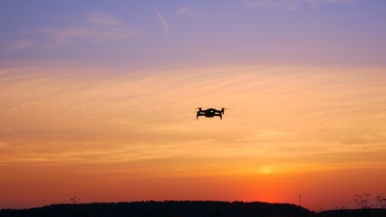 The drone moves from side to side in the bright sunset sky. The silhouette of a moving drone in flight with blinking lights against an orange sky flooded with sunset light. Concept of new technologies