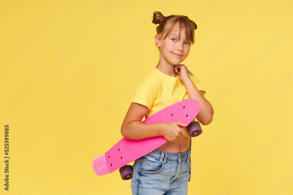 Wall mural A cute little girl 6 years old in a bright tulle skirt and a white t-shirt and stylish glasses holds a bright colored skateboard on a yellow background