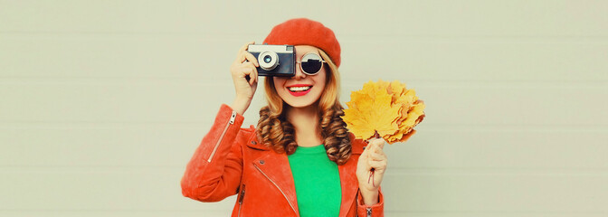 Autumn portrait of happy smiling young woman with film camera and yellow maple leaves wearing red french beret on gray background