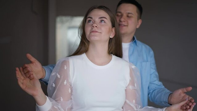 Portrait of excited young woman in new flat with man covering eyes with hands surprising partner with repair. Loving Caucasian couple indoors in apartment talking and smiling