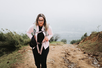 Tired young latin plus size woman with backpack on, climbing a hill and hiking in a beautiful...