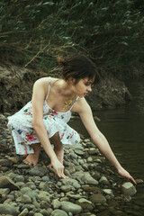 A girl in a dress on the river bank stretches her hand to the water. Walk in nature.