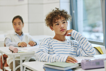 Portrait of curly hair young boy sitting at desk in classroom in minimal white setting, copy space