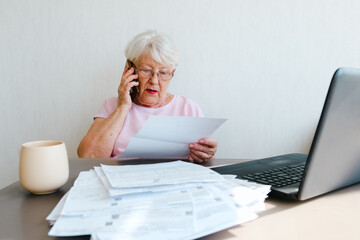Senior woman holding papers busy at laptop managing house utility bills or finances, aged female using computer working with bank loan or mortgage documents online. Elderly 