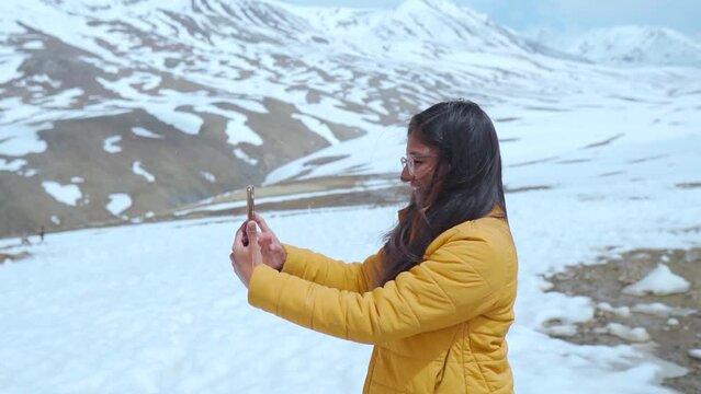 Portrait of young Indian female tourist taking photo-video using mobile phone during light snowfall at Baralacha La Pass, Himachal Pradesh, India. woman shooting video during snowfall at Mountains