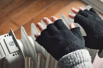 Close-up of a person's hands wearing a wool cardigan and black wooly fingerless gloves close to a domestic radiator trying to keep warm in the winter