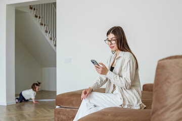 Brunette woman uses mobile phone, reads social networks, relaxes and sits on sofa. Baby is crawling
