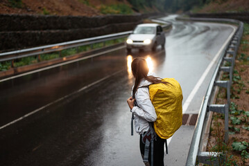 Hitch-hiker woman looking away while waiting cars with yellow backpack in nature