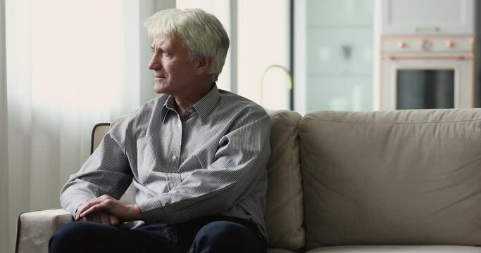 Unhappy depressed lonely elderly man sitting on couch at home, looking at window away in deep thoughts, thinking over loneliness, bad news, loss, gried. Elderly age problems concept