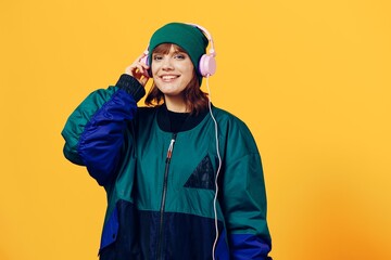 a happy, joyful woman in a green jacket and hat enjoys music standing in pink headphones on a yellow background, holding them with her hands and looking at the camera. Horizontal photo