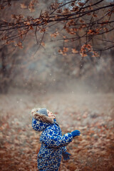 Fototapeta na wymiar A child, a boy in a warm jumpsuit stands in the park under a tree and looks at the first snow falling, the beginning of winter or the end of autumn, late autumn